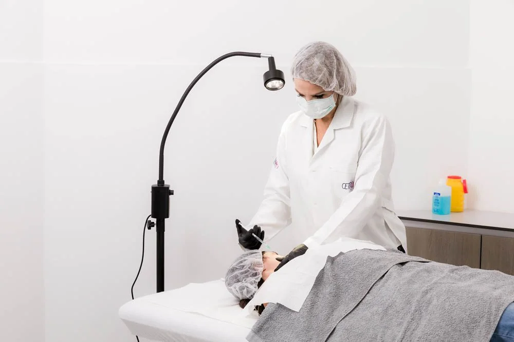 Profissional de saúde realizando procedimento estético ou dermatológico em paciente em uma clínica, com luz de exame e equipamentos de higiene ao fundo.