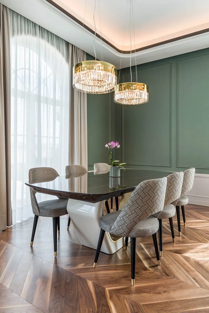 Sala de jantar moderna com mesa de vidro escuro, cadeiras estofadas em tecido com diferentes padrões, cortinas brancas e paredes verdes, iluminação de dois lustres dourados com cristais e piso de madeira em estilo parquet.