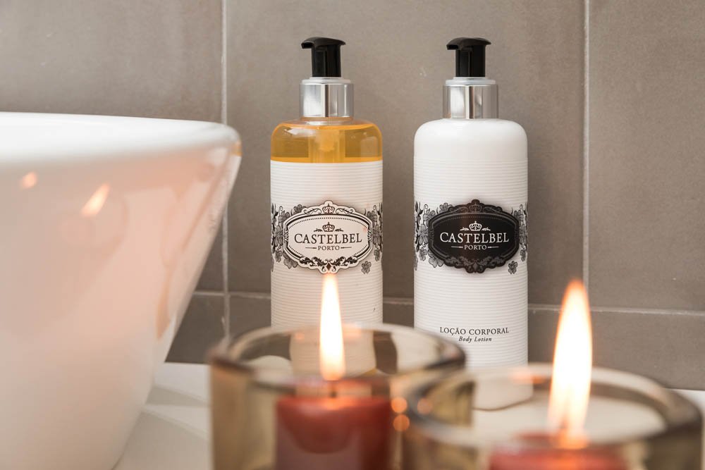 Velas acesas na frente de duas garrafas de hidratante, uma de óleo e outra de loção corporal, na varanda de um banheiro com paredes de azulejo.