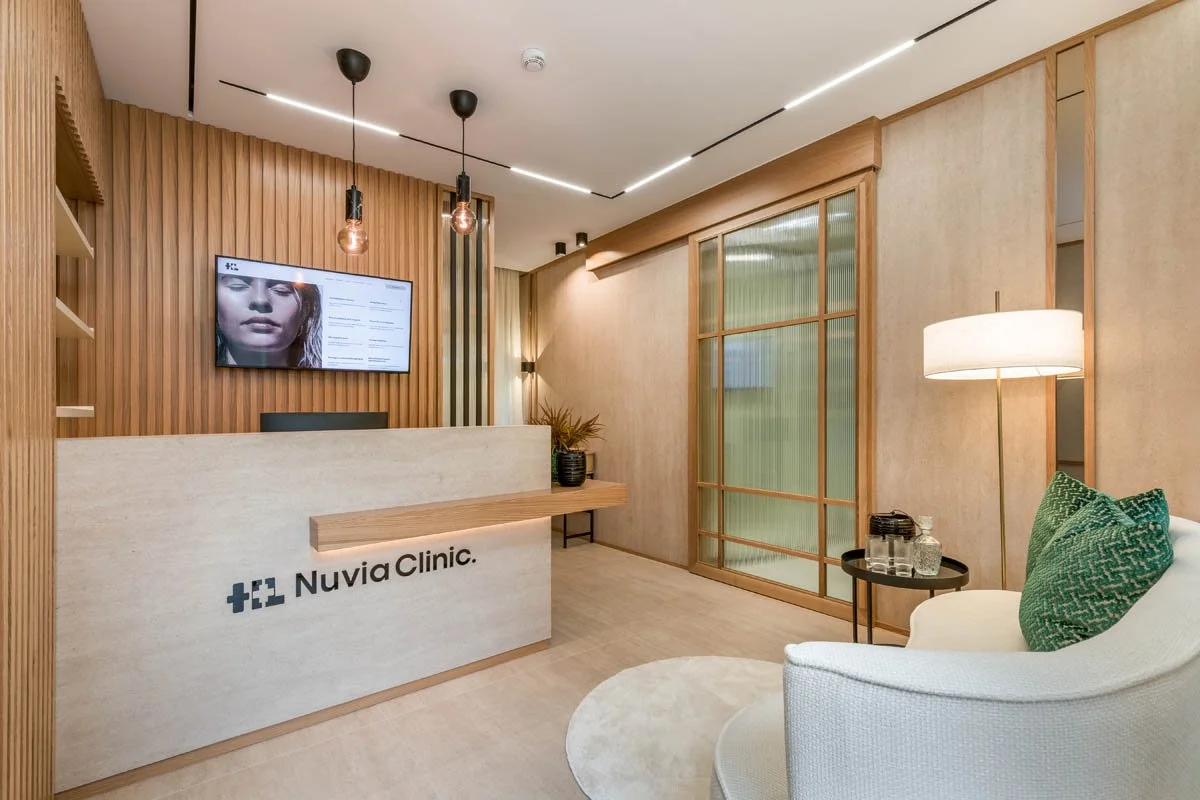 Recepção de clínica moderna com painel de madeira, televisão na parede, móveis de ambiente neutro com detalhes em verde, cadeira de descanso branca com almofadas verdes, piso de madeira clara, iluminação suave e ventilação natural