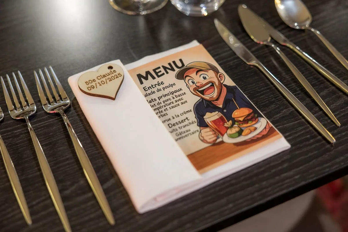 Menu de restaurante ao lado de talheres em mesa de jantar. Na frente, uma lembrança de coração de madeira com a inscrição '50e Claudé 09/10/2025'.