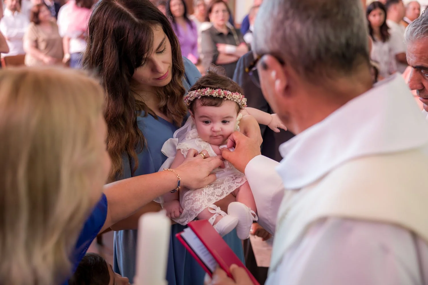 Bebê sendo batizada por um padre, com familiares ao redor em uma cerimônia religiosa.