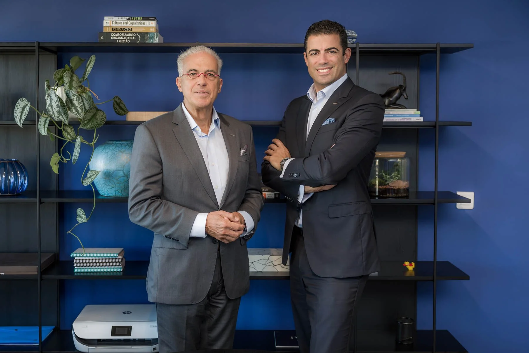 Dois homens de terno estão posando para a foto em frente a uma estante com livros, plantas e objetos decorativos. Um deles é mais velho e usa óculos vermelhos, o outro é mais jovem com cabelo escuro.