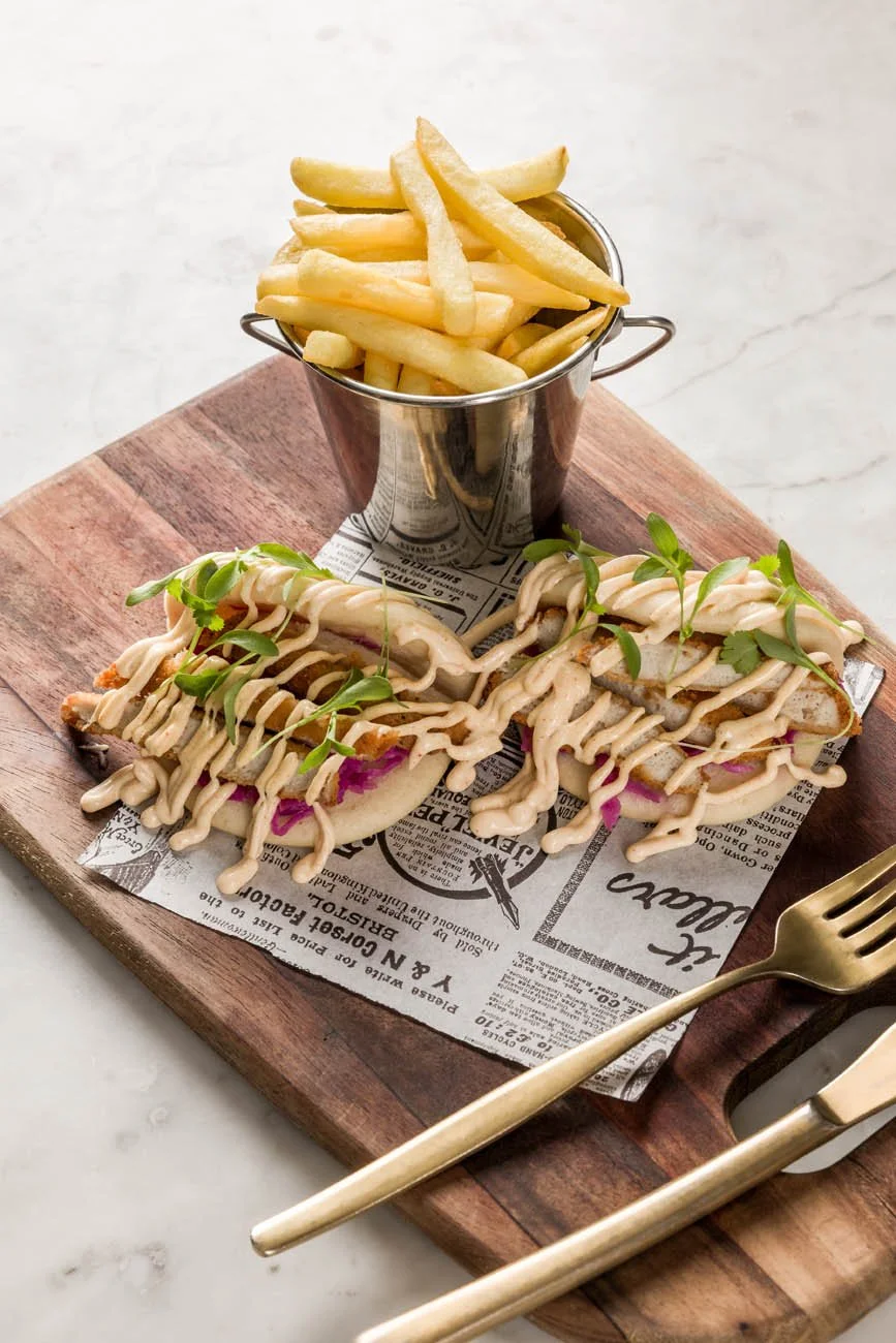 Sanduíche de pão ciabatta com carne, alface, cebola roxa, molho e gérberas, acompanhado de uma porção de batatas fritas em um recipiente de metal, sobre uma tábua de madeira com papel decorativo e garfo dourado ao lado.