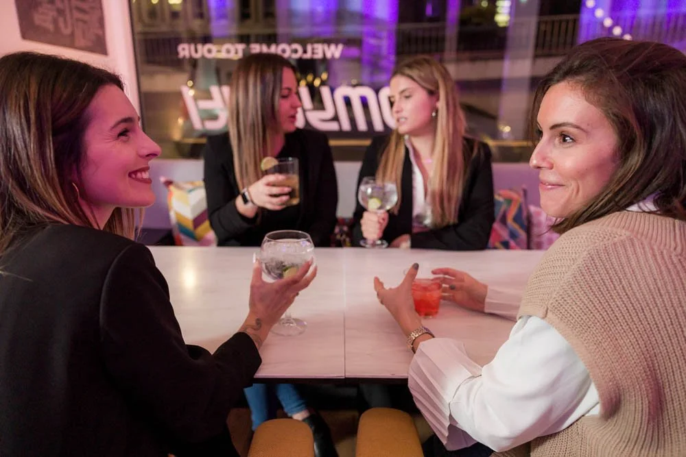Quatro mulheres conversando e sorrindo em um bar ou restaurante, cada uma segurando um drink.