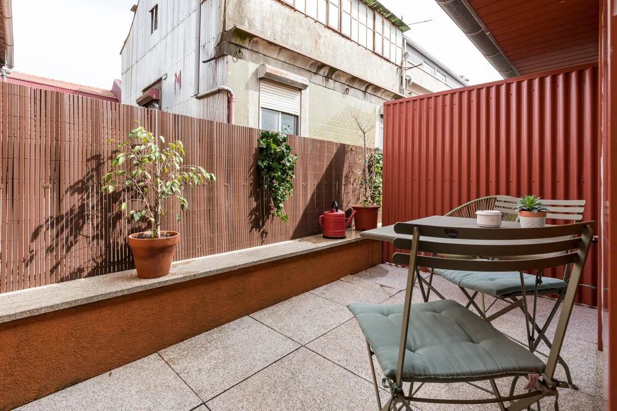 Varanda com mesa de metal com duas cadeiras acolchoadas, uma suculenta em vaso, um regador vermelho e uma planta em vaso na parede de cerca de madeira