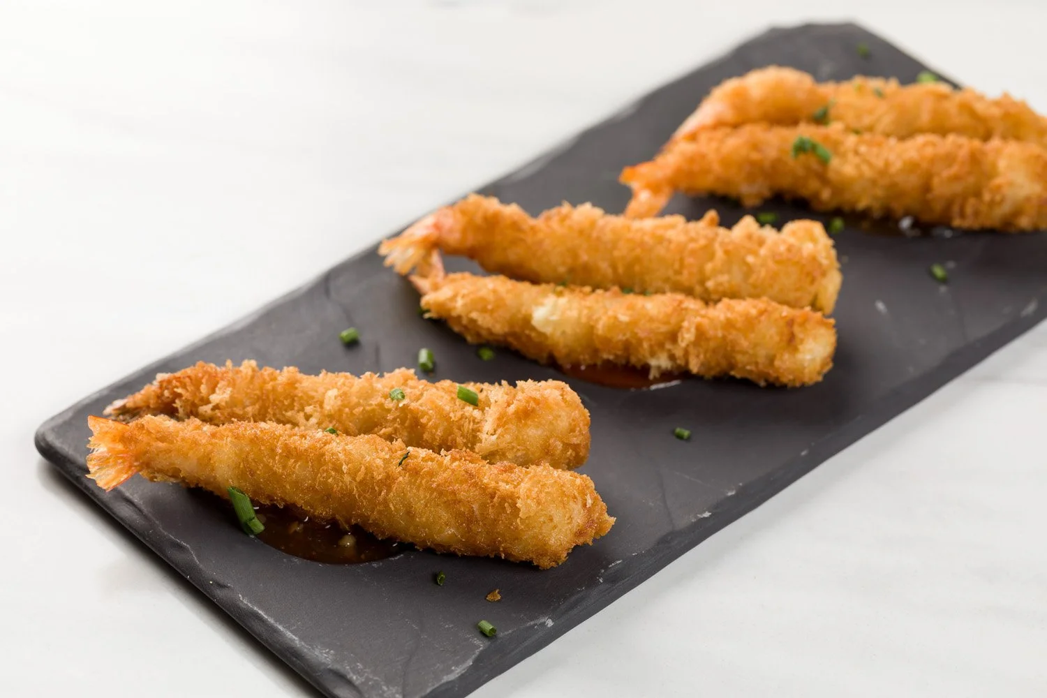 Porção de filés de peixe empanados e fritos, decorados com cebolinha, sobre uma placa preta