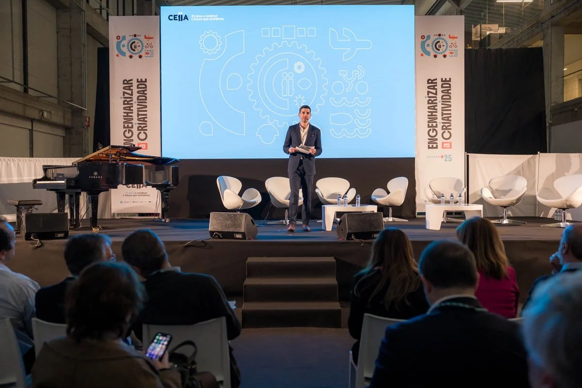 Homem de ternojavaxapresentando em palco durante conferência ou evento, com cadeiras e um piano ao lado, tela de projeção ao fundo e público assistindo.