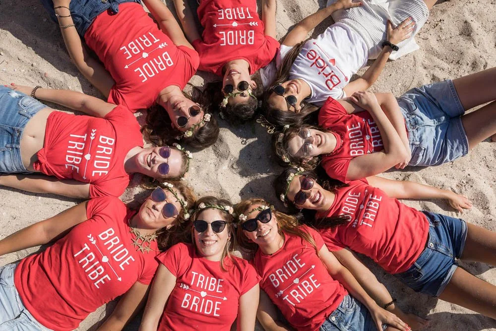 Grupo de mulheres deitada na areia de praia formando um círculo, usando camisetas vermelhas com frases relacionadas a casamento, como "Bride Tribe" e "Bride". Todas usam óculos escuros e estão sorrindo, com alguns decorados com coroas de flores na ca