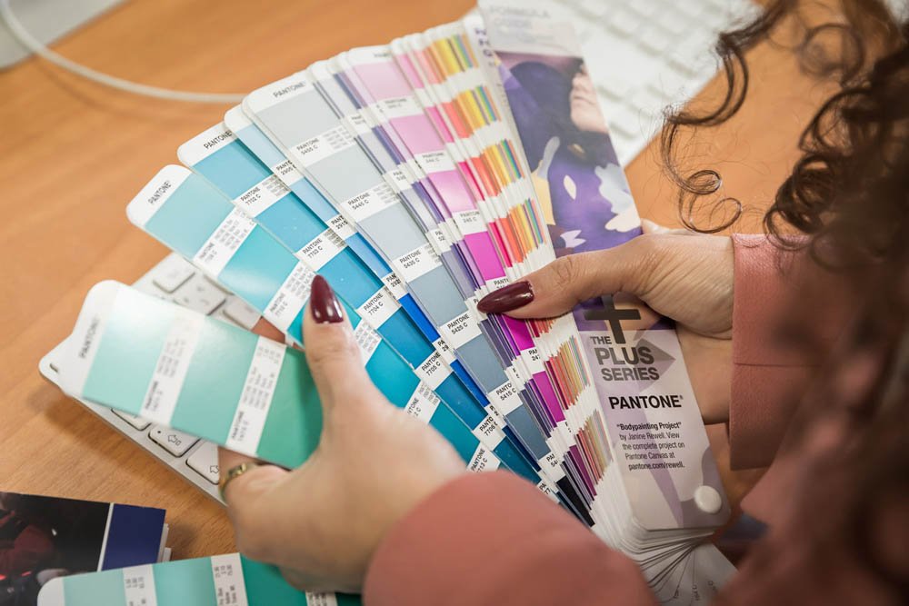 Pessoa segurando amostras de cores Pantone, organizada em fanfarras, com uma mesa de computador ao fundo.