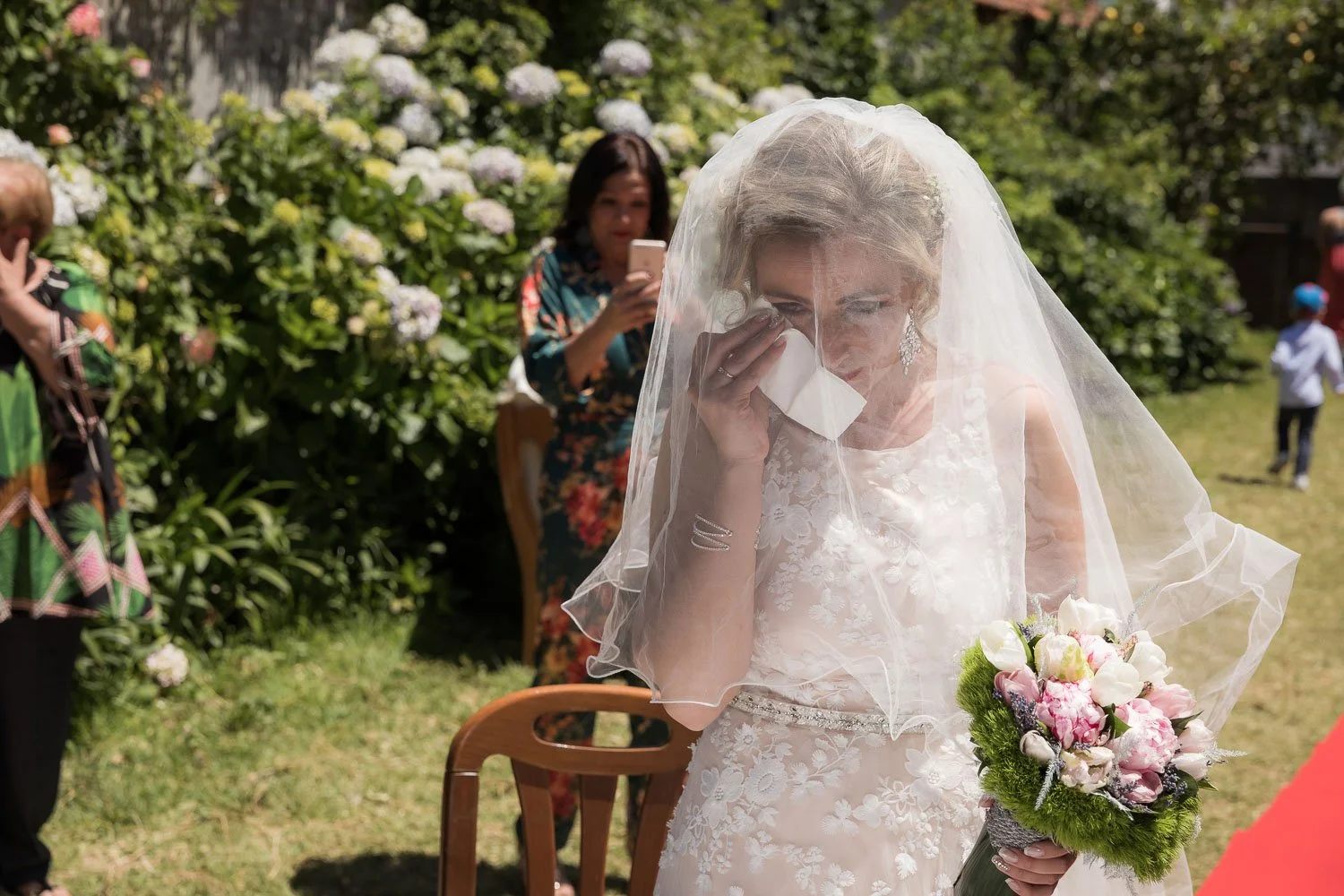 Noivo(a) chorando com uma toalha de papel na mão, vestindo vestido de casamento, segurando buquê de flores, ao ar livre com vegetação ao fundo, enquanto uma mulher ao fundo tira foto com celular.