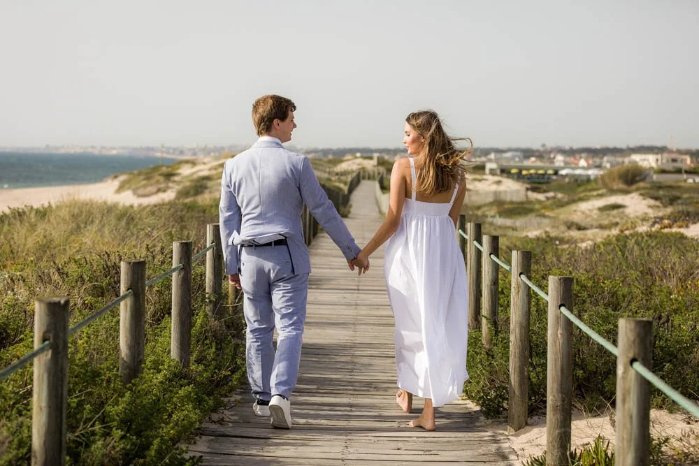 Casal andando de mãos dadas por passarela de madeira na praia, com vegetação ao redor e mar ao fundo.