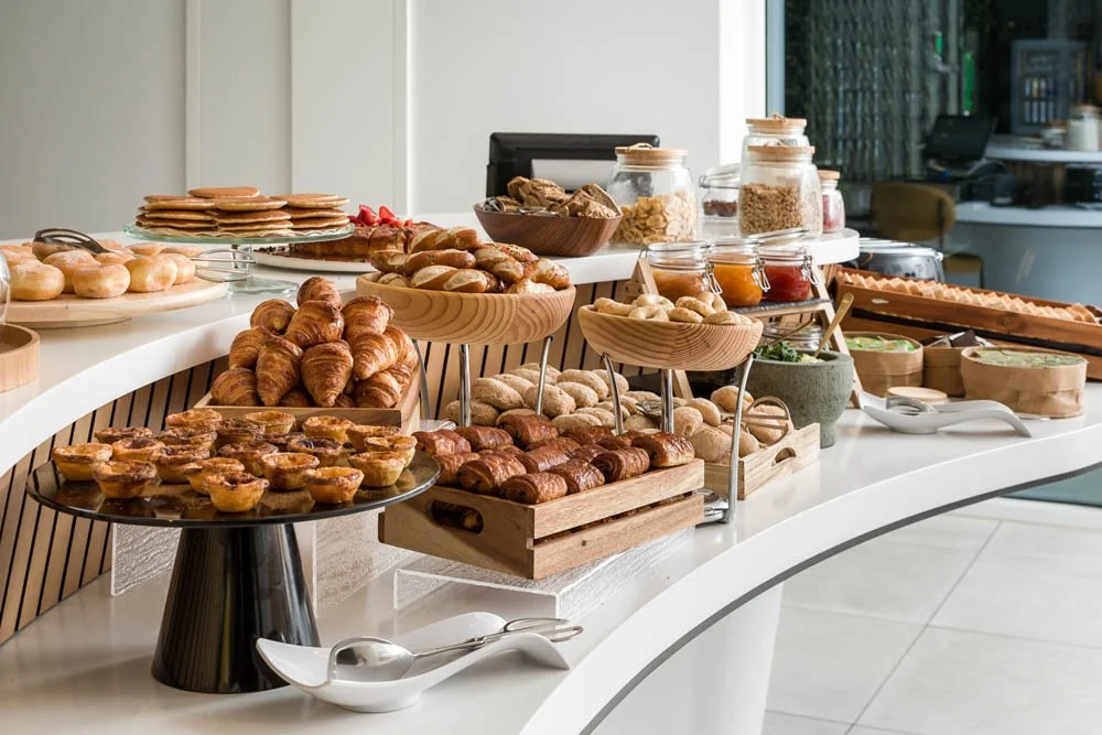 Buffet de café da manhã com pães, croissants, geleias, ovos e outros alimentos em uma mesa moderna.
