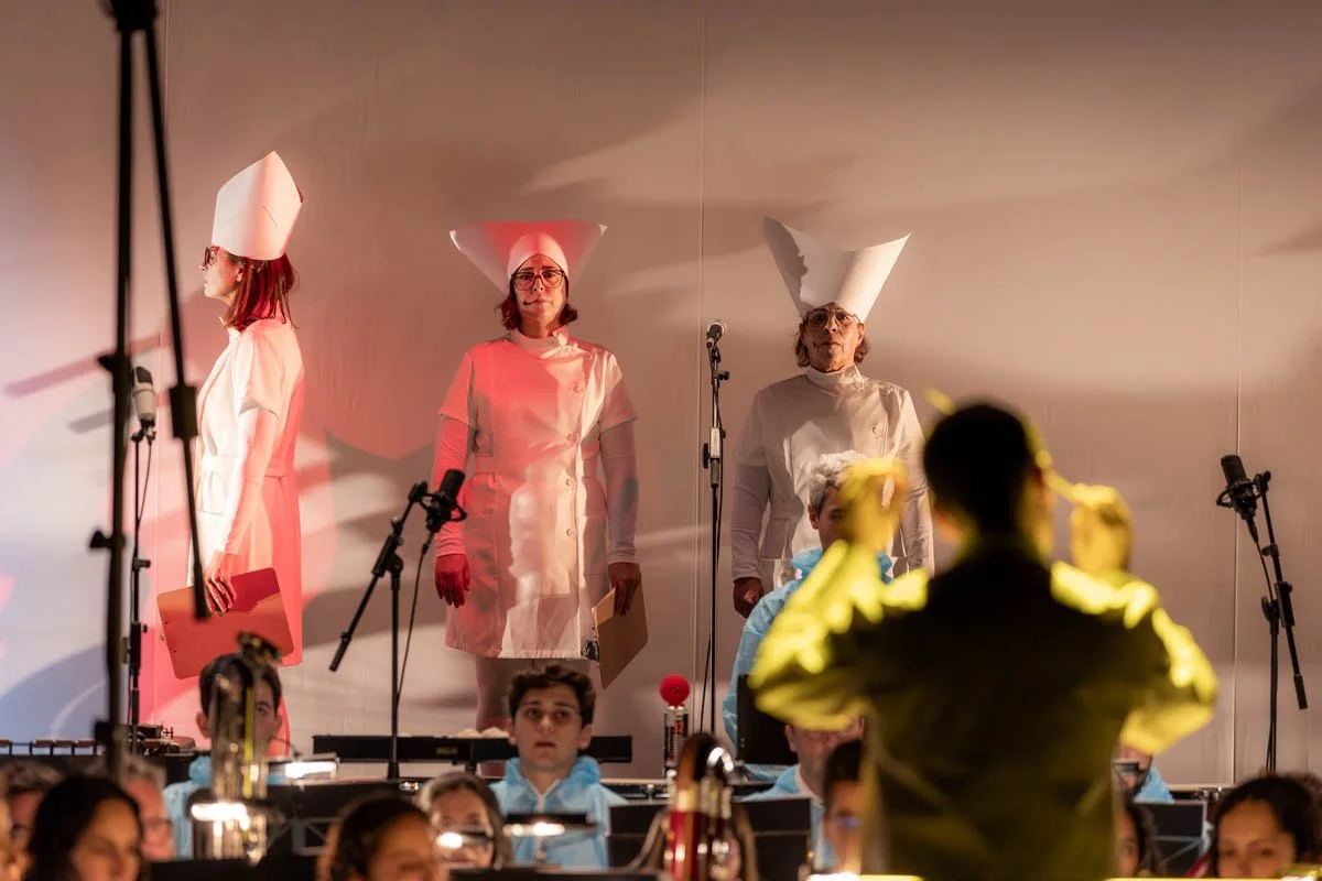 Cenário de show com três mulheres na frente, vestidas com jalecos brancos e chapéus de chef, em um palco com microfones. Relações de luz e uma orquestra visível na parte inferior da imagem.