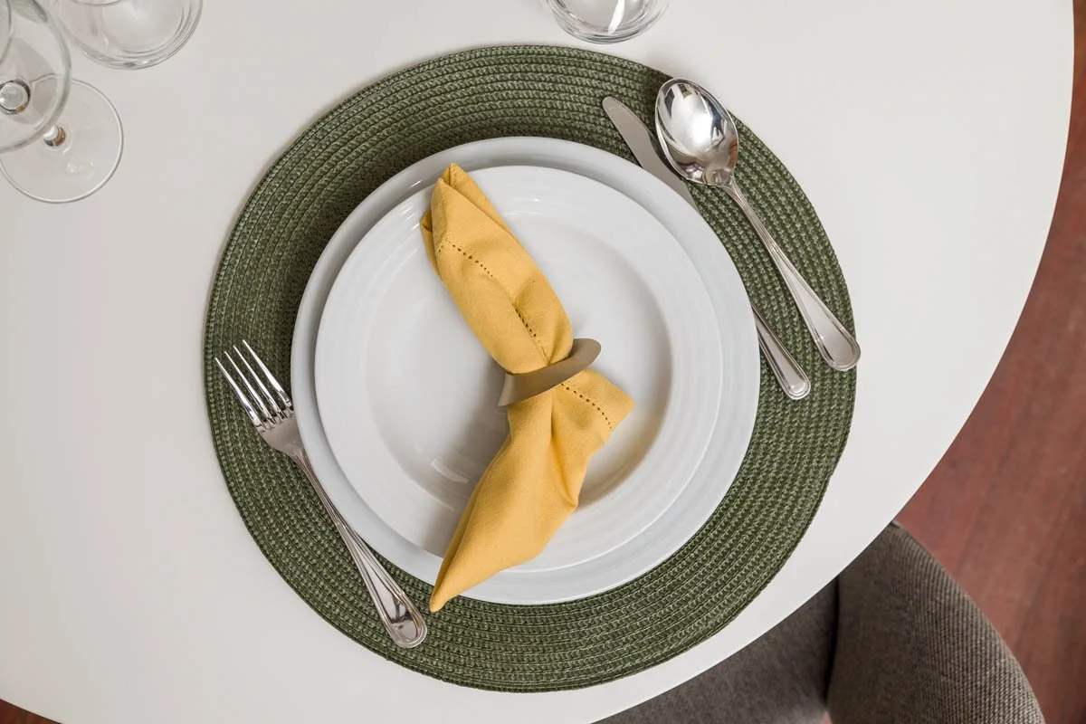 Prato branco com talheres ao redor, guardanapo amarelho amarrado com tampinha de plástico, sobre uma toalha verde escura em uma mesa