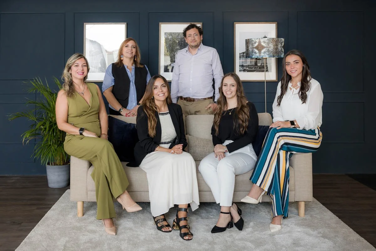 Grupo de sete pessoas posando para uma foto em uma sala com parede azul escura, quadros na parede, uma planta ao lado e uma luminária. As pessoas estão sentadas e em pé, todas sorrindo.