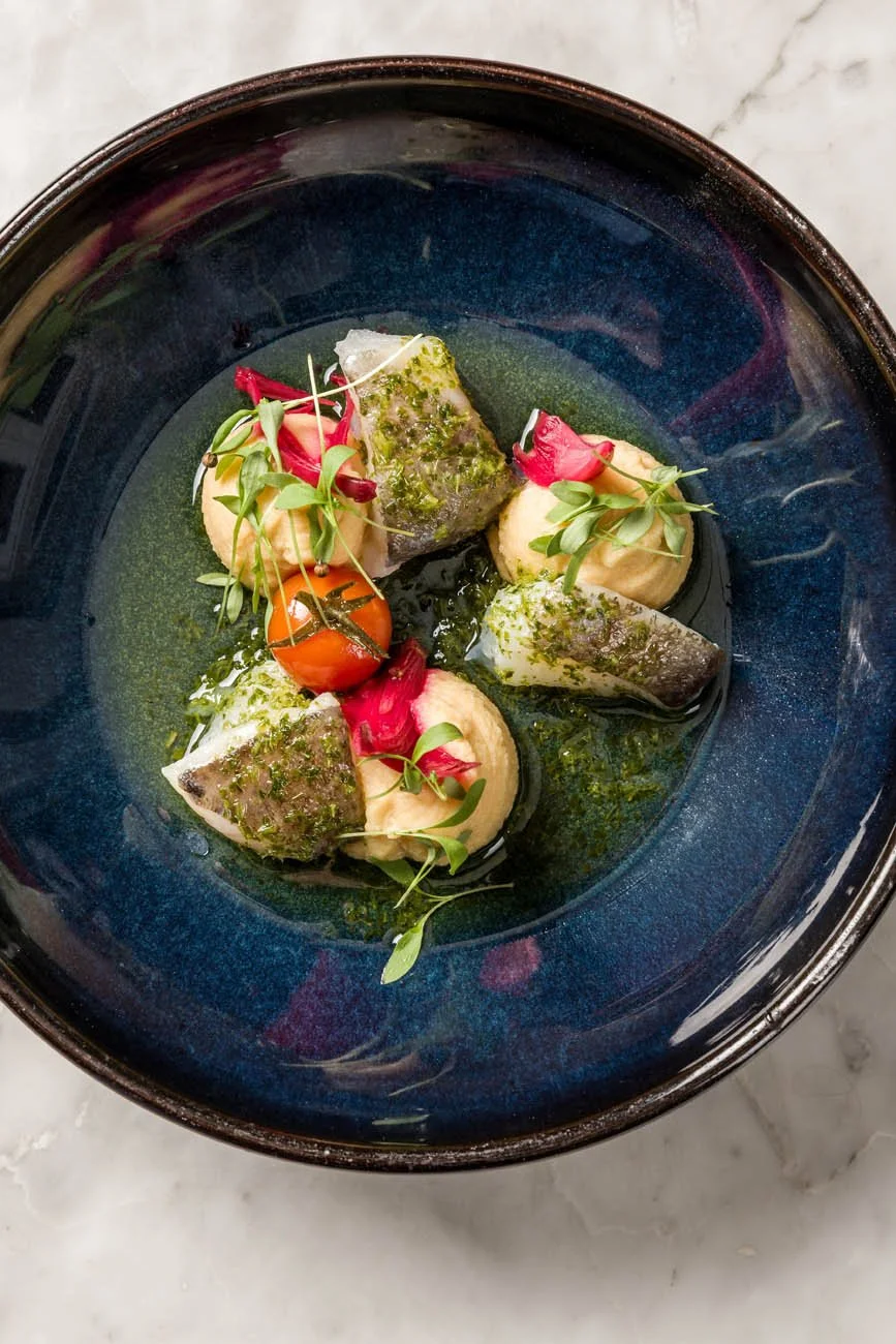 Prato de apresentação sofisticada com rolinhos de peixe, tomate cereja, purê e microverdes, decorado com flores comestíveis e molho verde.