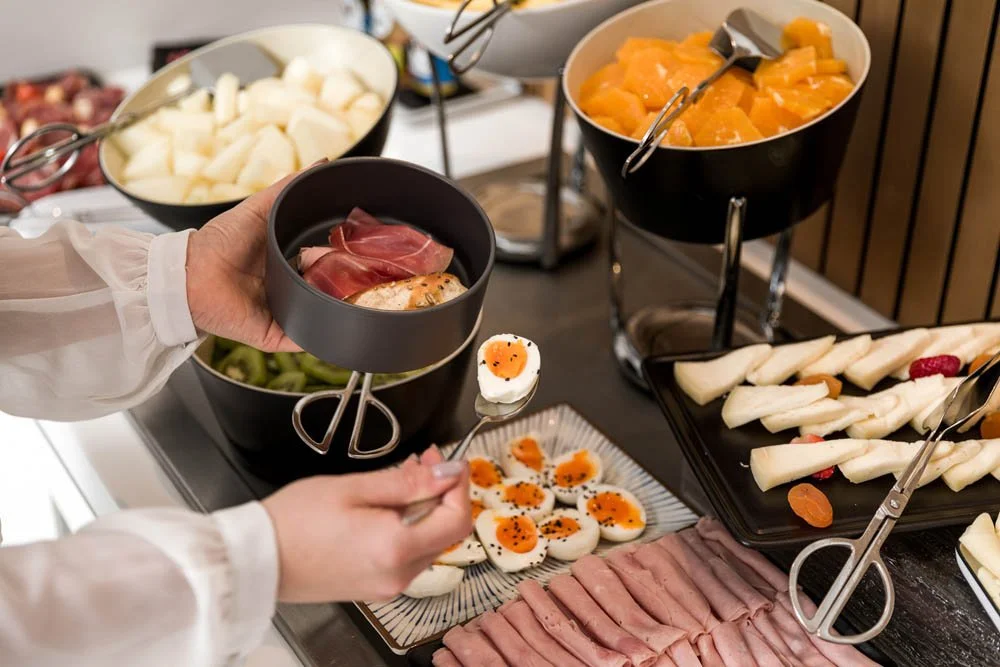 Mesa com alimentos em estilo buffet, incluindo frutas, ovos cozidos, presunto, queijo, melão, melão cortado, e outros itens de café da manhã.