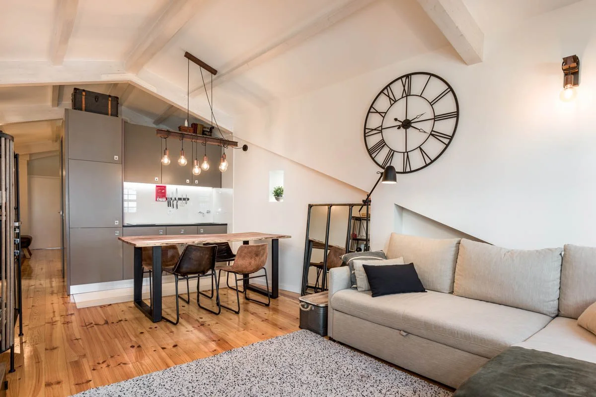 Sala de estar com sofá bege, almofadas pretas e cinza, relógio de parede grande de ferro, iluminação de teto e parede, e uma área de cozinha com mesa de madeira e cadeiras.