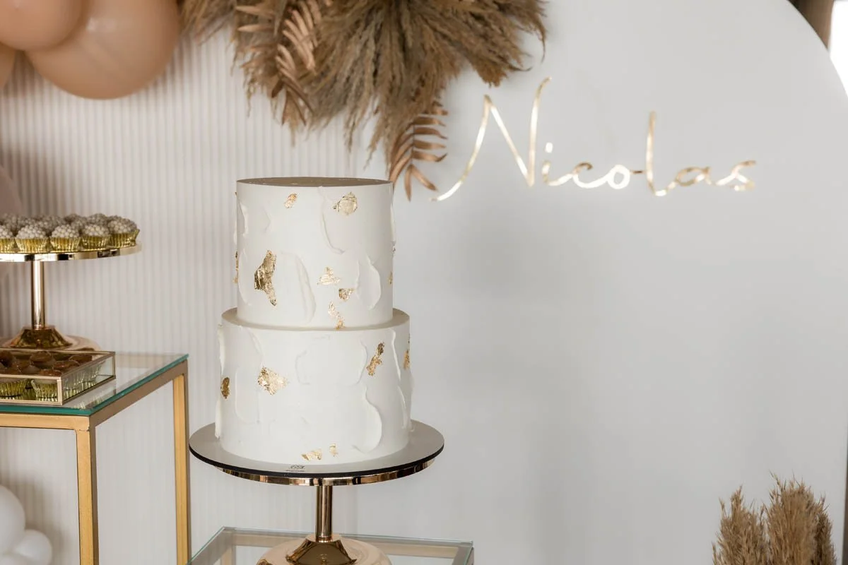 Bolo de aniversário branco de dois andares com detalhes dourados, decorando uma mesa de festa com nome 'Nicolas' ao fundo.