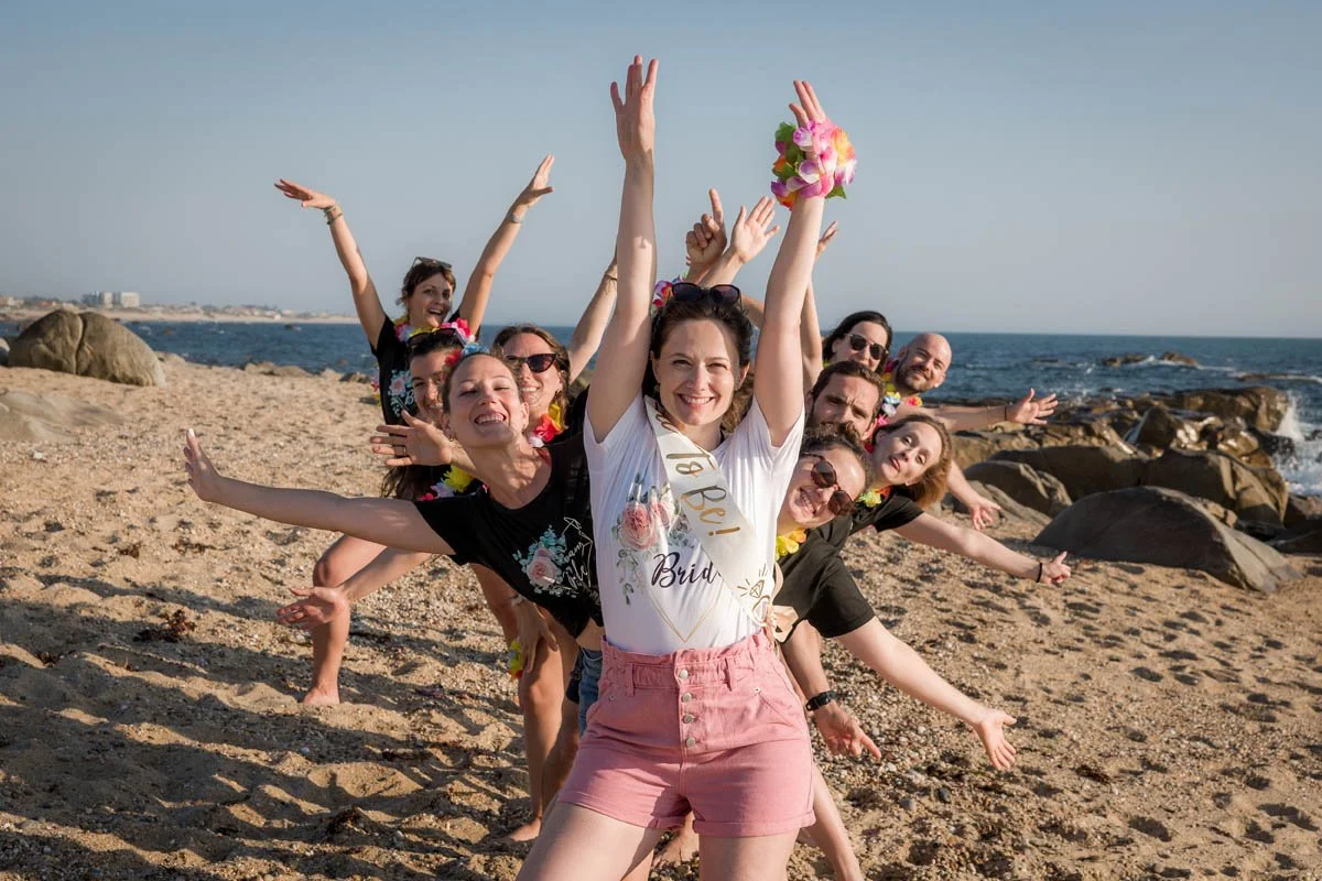 Grupo de pessoas celebrando na praia com sorriso, roupas coloridas e acessórios de festa, com o mar ao fundo.