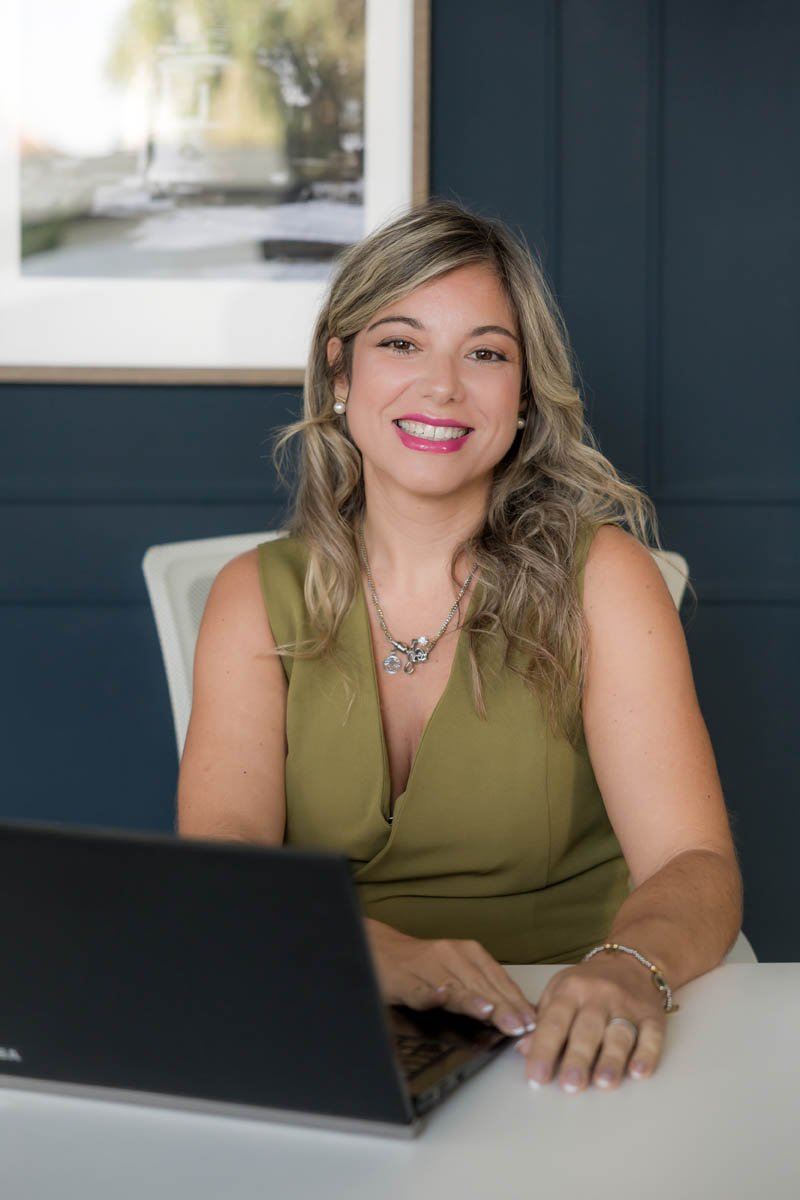 Mulher sorridente com cabelos loiros ondulados, usando um vestido verde, brincos de pérola, colar de corrente com pingente, sentada em uma mesa de escritório com laptop, fundo azul e janela ao fundo com vista para árvores.