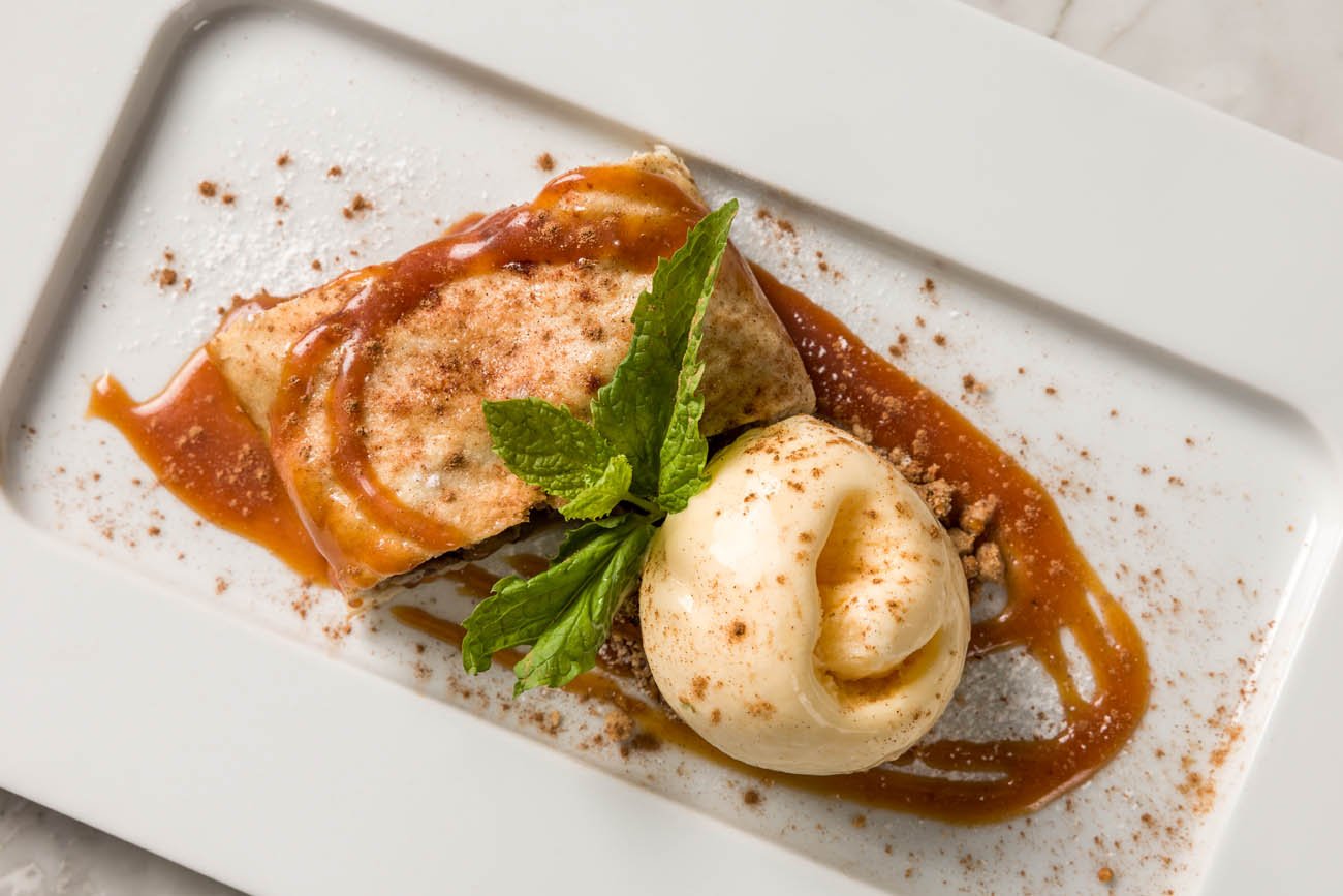 Sobremesa com fatia de cheesecake, sorvete, folhagem de hortelã, calda de caramelo, e pó de cacau sobre prato branco.