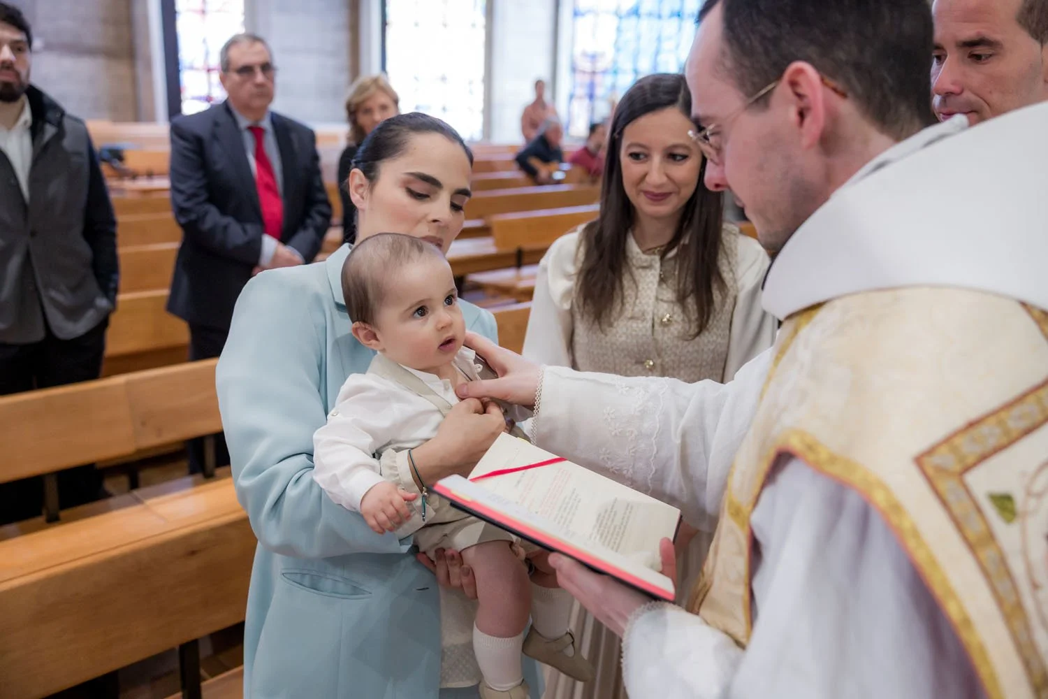 Bebê sendo batizado por padre durante cerimônia religiosa na igreja, com familiares observando ao fundo.