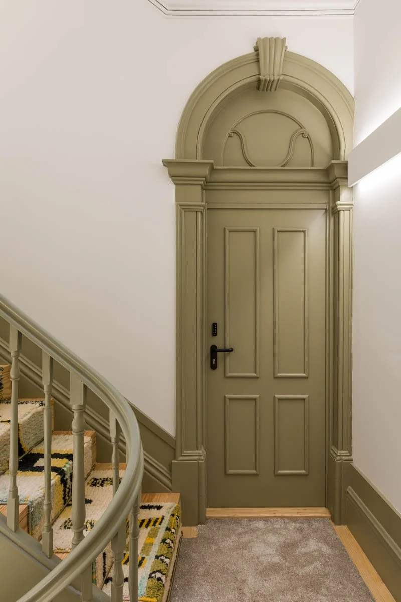 Porta clássica de madeira com detalhes ornamentais, situada na parede de uma escada com tapete colorido.