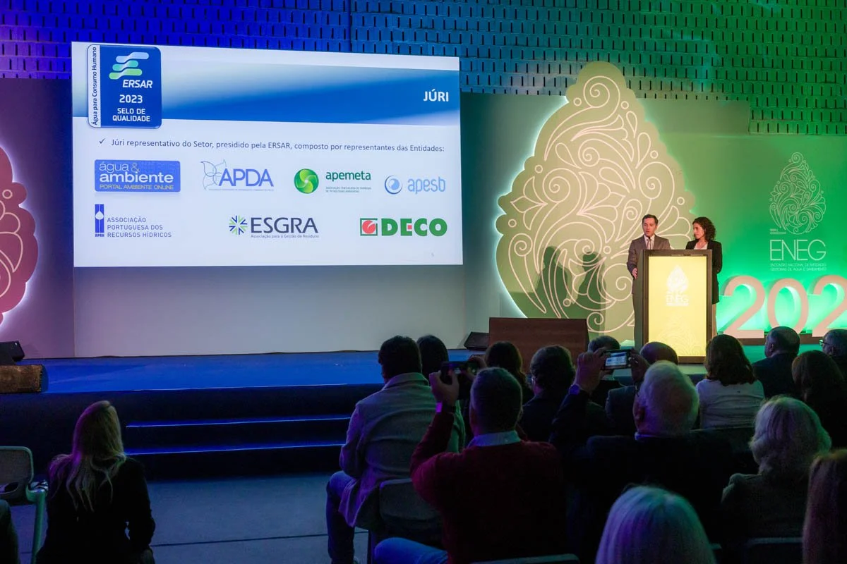 Apresentação em um evento do setor de recursos hídricos, com painel digital mostrando o logo do ERSAR e nomes de entidades relacionadas ao setor, em um palco iluminado com duas pessoas falando no púlpito, e uma audiência assistindo.