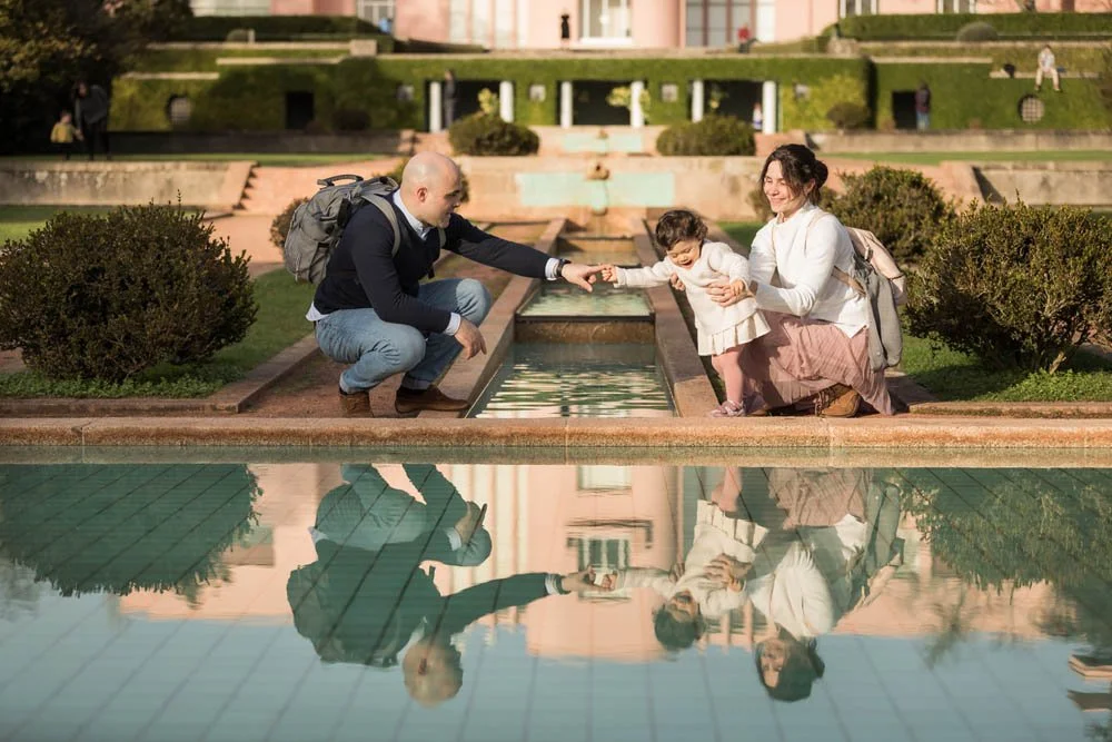 Família com pai, mãe e filha na beira de uma fonte em um parque, brincando e se divertindo.