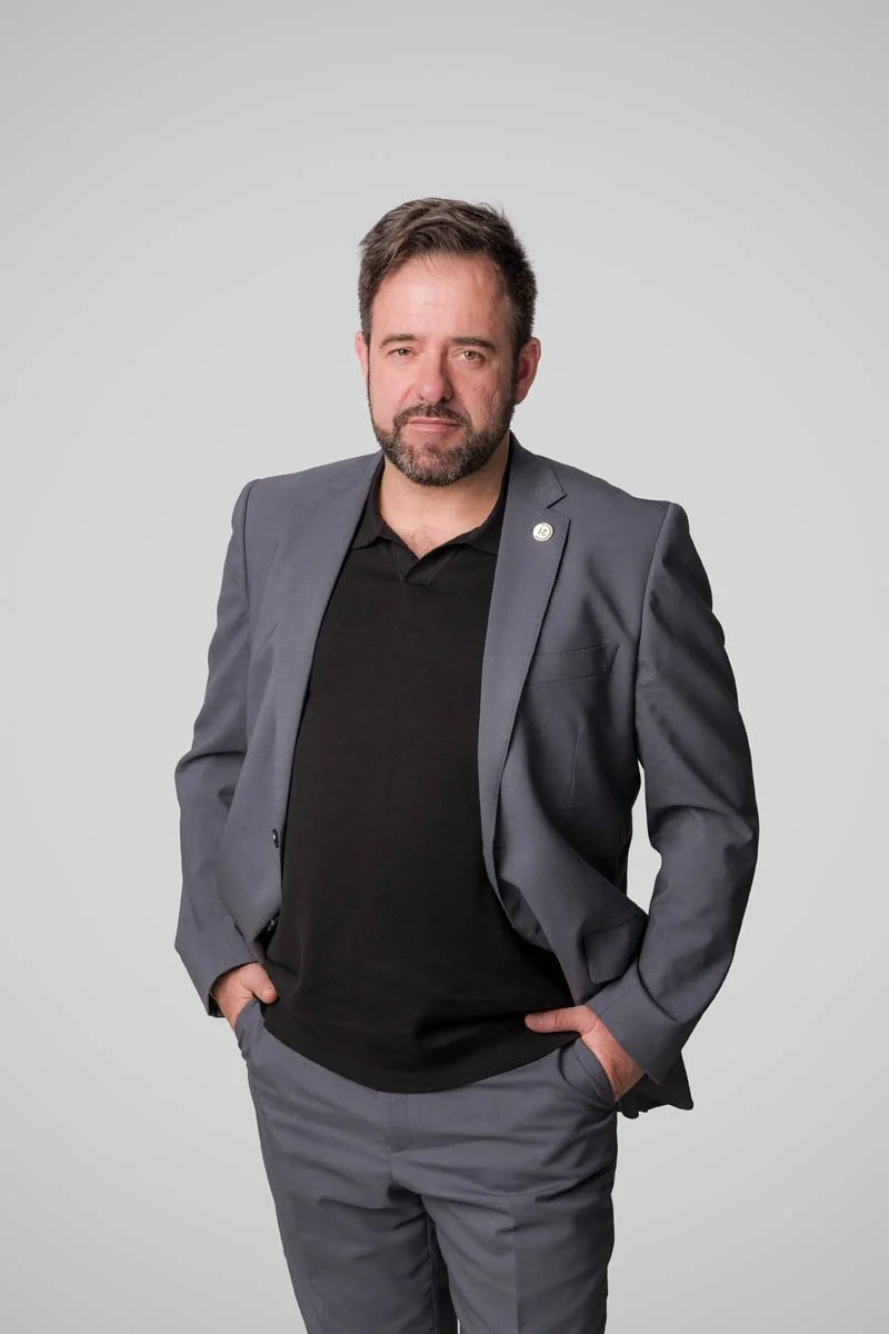 Homem com barba, vestindo terno cinza escuro e camiseta preta, posando com as mãos nos bolsos contra fundo cinza claro.