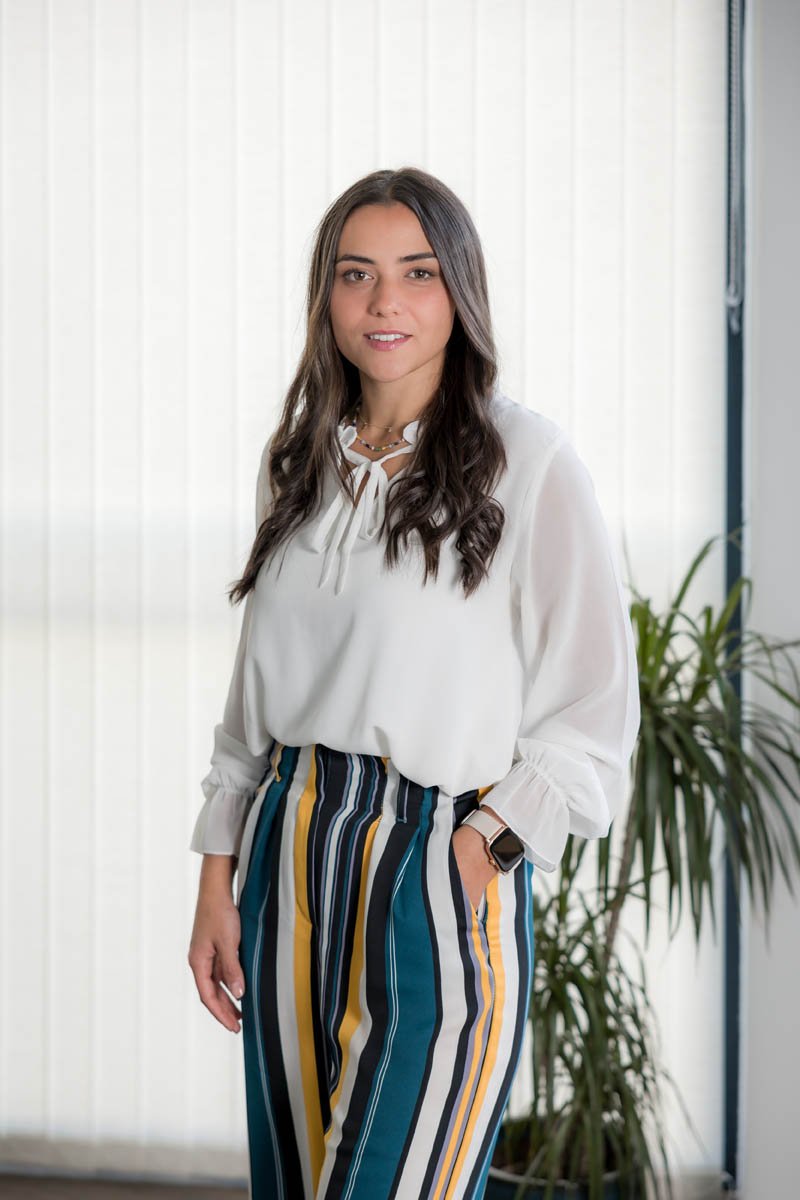Mulher com cabelo castanho escuro, vestido branco de mangas longas com laço no pescoço, calça de riscas coloridas, na frente de uma porta de vidro com persianas brancas ao fundo, segurando a mão na cintura, com planta ao lado.
