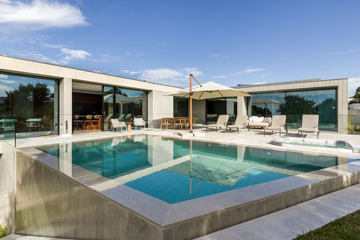 Casa moderna com piscina e área de convivência ao ar livre, incluindo espreguiçadeiras, mesa de jantar com guarda-sol e janelas de vidro.
