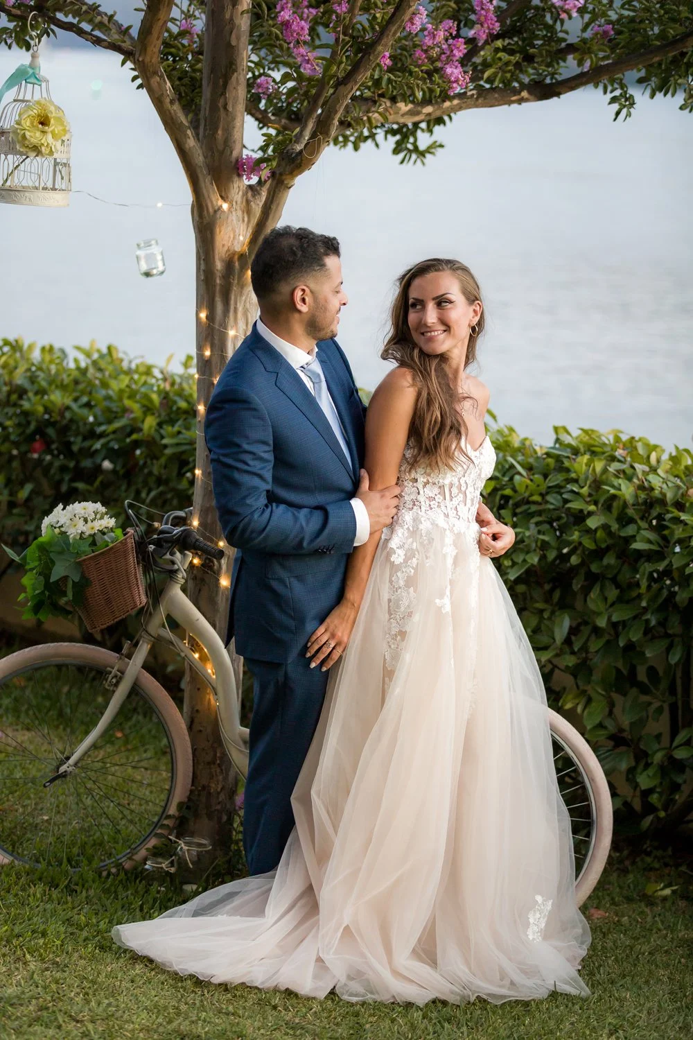 Casal de noivos ao ar livre, na frente de uma árvore decorada com luzes e flores, com uma bicicleta decorada com flores ao lado, durante o pôr do sol ou o entardecer.