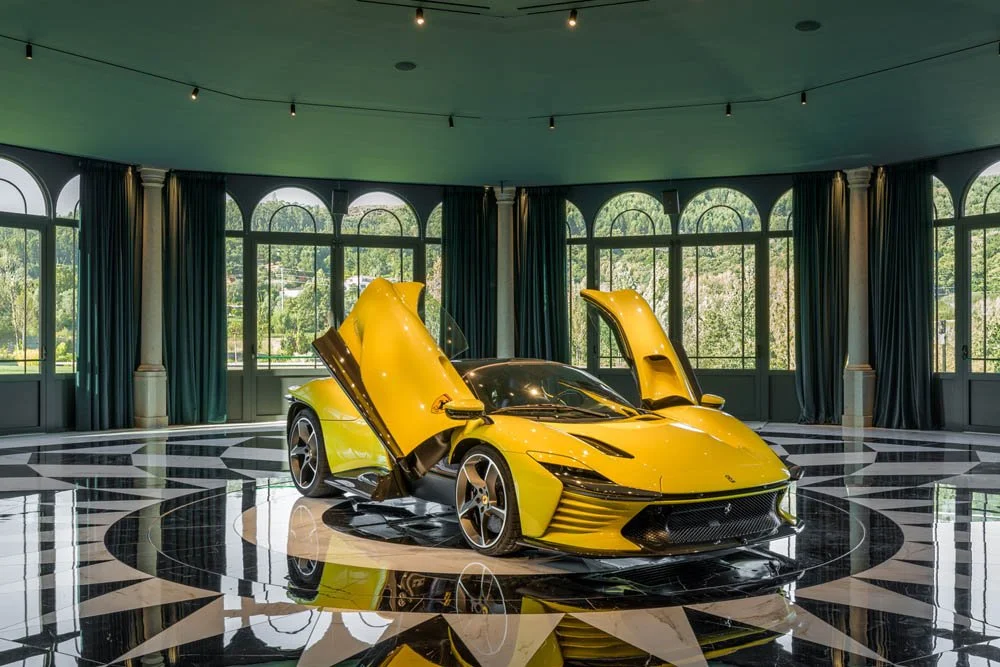 Carro esportivo amarelo com portas elevatórias abertas, exibido em sala com piso preto e branco e janelas grandes com cortinas verdes.