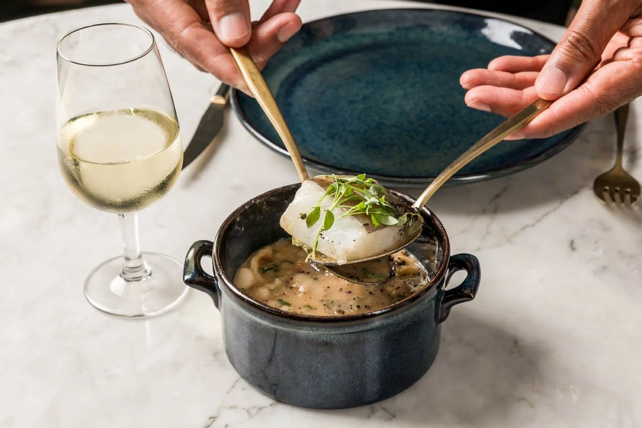 Pessoa colocando peixe cozido com ervas em prato de barro, ao lado de taça de espumante em mesa de mármore.
