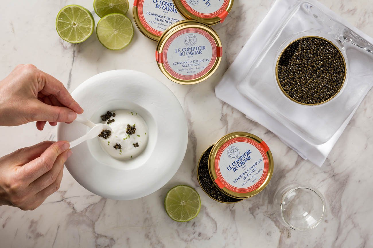 Pessoa usando uma colher para colocar ovas de capricórnio em uma tigela de cerâmica branca, com limões cortados ao redor e latas de ovas de capricórnio ao lado, em uma superfície de mármore.