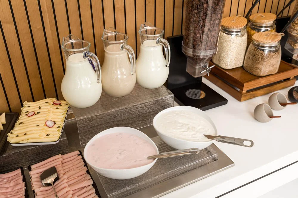 Opções de iogurte ou sobremesas em tigelas brancas, várias jarras de leite, e dispensadores de cereais, em uma estação de café da manhã.