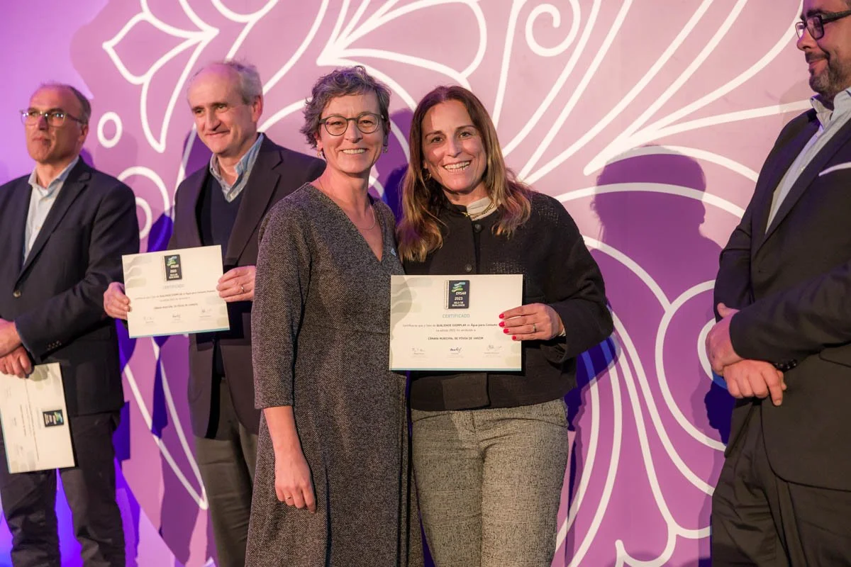 Duas mulheres sorrindo, uma delas segurando um certificado, no palco durante uma cerimônia de premiação. Ao fundo, outras pessoas também segurando certificados, todos de terno, com uma decoração floral dourada.
