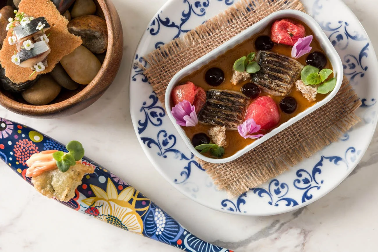 Prato com peixe, frutas e flores, acompanhamentos em tigelas decoradas com detalhes azuis, sobre uma mesa de mármore