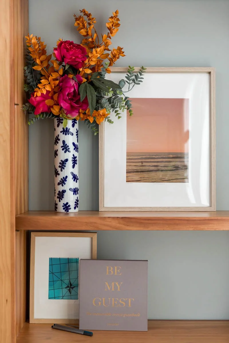 Vaso com flores coloridas, quadro de praia com pôr do sol, quadro com mensagem "Seja meu convidado" e uma caneta em uma estante de madeira.