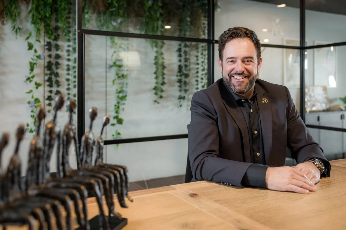 Homem sorridente sentado em uma mesa de madeira com uma peça de troféu ao seu lado em um ambiente moderno com decoração de plantas.