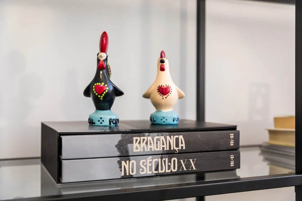 Duas estatuetas coloridas de formato de galinha em cima de dois livros empilhados, com um fundo de prateleira e objetos ao lado.