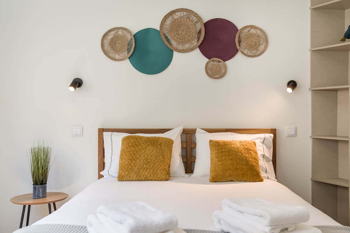 Quarto com cama de madeira, duas almofadas amarelas, paredes decoradas com cestos de vime, luminárias ao lado da cama, e uma prateleira do lado direito