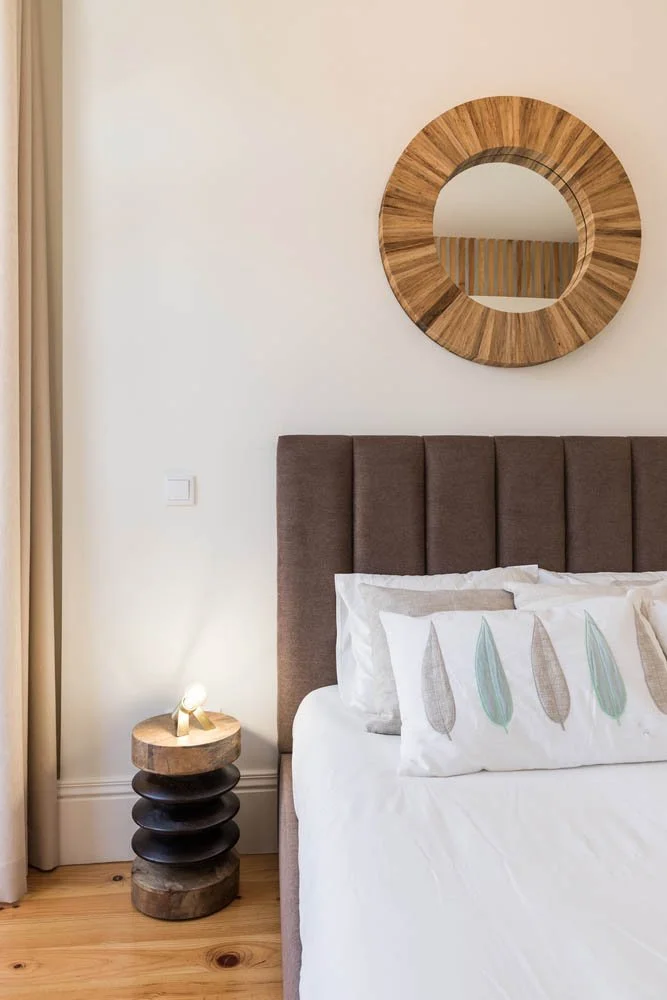 Quarto com cama de cabeceira acolchoada, travesseiros decorativos com plumas em cores suaves, espelho redondo de madeira na parede e luminária de cabeceira em cima de uma mesa de madeira com detalhes escuros, ao lado de uma cortina bege.