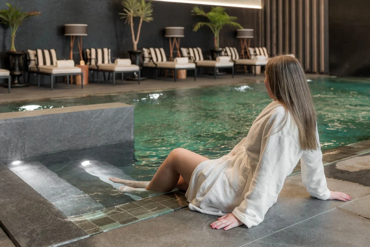 Mulher com cabelo castanho claro usando roupão branco, sentada na borda de uma piscina interior, com as pernas na água, em um ambiente moderno com cadeiras e plantas ao fundo.