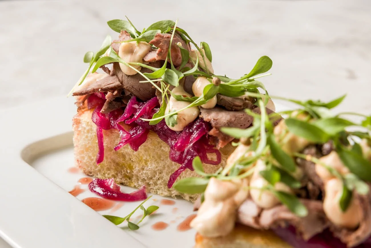 Torrada de pão com carne, molho rosa, brotos verdes e cebolas em conserva, decorada com gotas de molho.