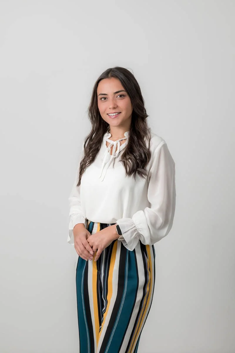 Mulher com cabelo castanho escuro, vestindo blusa branca e calças listradas em azul, amarelo, branco e preto, sorrindo, contra fundo branco.