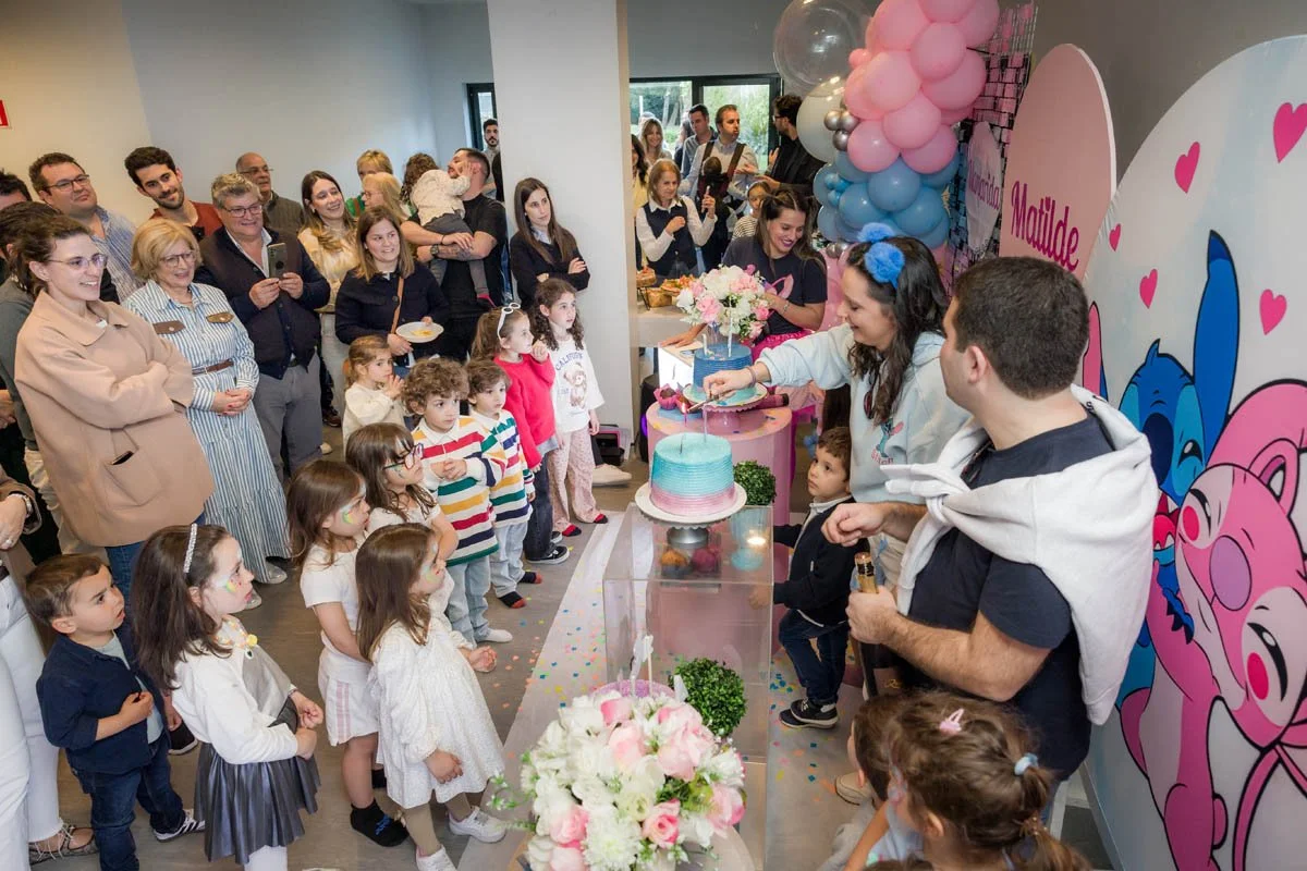 Crianças e adultos assistindo a uma festinha de aniversário infantil com decoração pink e azul, balões, papelaria temática, bolo colorido, e uma mulher cortando o bolo enquanto uma mulher com aurora azul na cabeça entrega uma fatia às crianças.